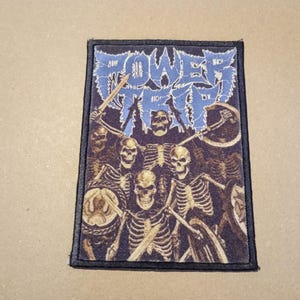 Puede incluir: Parche bordado en blanco y negro con el texto "POWER TRIP" en una fuente azul y blanca. El parche presenta un grupo de esqueletos sosteniendo espadas y escudos.