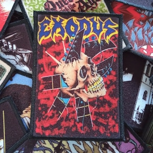 May include: A black and red embroidered patch with the word "EXODUS" in blue and white letters. The patch features a skull with a guitar neck piercing through it.