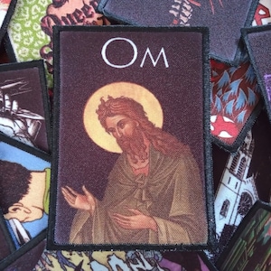 Può includere: Una toppa ricamata nera e marrone con l'immagine di un uomo barbuto con un'aureola. Il testo "OM" è sopra l'immagine.