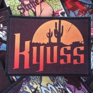 May include: A black and orange embroidered patch with the word "KYUSS" in a stylised font. The patch features a sunset with two cacti in the foreground.