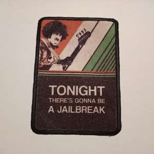 Può includere: Un cerotto in tessuto marrone e nero con un'immagine retrò di una persona che suona la chitarra. Il testo "TONIGHT THERE'S GONNA BE A JAILBREAK" è stampato sul cerotto.