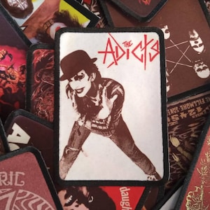 May include: A white and red embroidered patch with a black border. The patch features a black and white image of a person wearing a black and white striped shirt and a black bowler hat. The text "The Adicts" is in red and white letters.