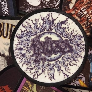 Puede incluir: Un parche redondo con la palabra "Kyuss" en morado, rodeado por un diseño blanco y morado. El parche tiene un borde negro y está sobre un fondo blanco. Otros parches son visibles en el fondo.