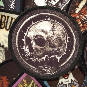 Könnte beinhalten: Ein schwarz-weißes Totenkopf-Patch mit weißem Hintergrund und einem schwarzen Totenkopf. Der Totenkopf ist von einem schwarzen Kreis mit weißem Rand umgeben. Das Patch ist auf einen schwarzen Stoff genäht.