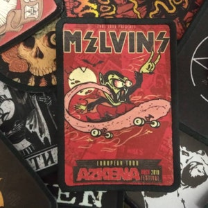 Può includere: Un patch ricamato rosso, bianco e nero con il testo "MZLVINS" in bianco. Il patch presenta un vampiro dei cartoni animati che fa skateboard su una lingua rosa. Il testo "EUROPEAN TOUR" e "AZKENA ROCK 2019 FESTIVAL" è stampato sotto l'immagine.