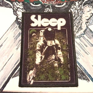 May include: A black and white embroidered patch with the word "Sleep" in white lettering. The patch features an astronaut in a dark spacesuit walking through a field of green plants.