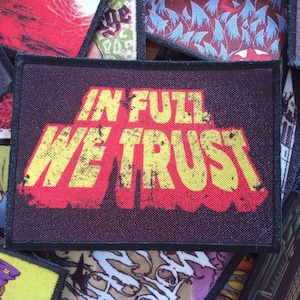 Peut inclure: Un patch noir avec un graphisme détérioré rouge et jaune qui dit "In Full We Trust".
