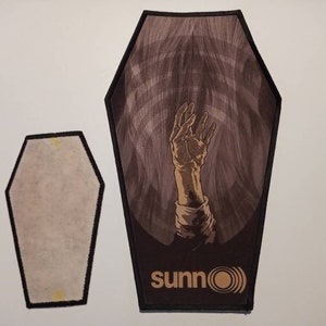 May include: Two black and brown coffin-shaped patches. The larger patch has a hand reaching up from a dark background with the word "sunn0" and a circle with a wave in it. The smaller patch is blank.