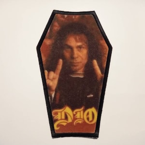 May include: Black and yellow coffin-shaped patch with a photo of Ronnie James Dio, the singer of the heavy metal band Dio, giving the rock and roll hand gesture. The text "DIO" is printed in yellow on the patch.