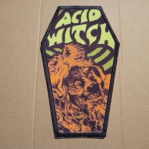May include: A black and orange coffin-shaped patch with the text "Acid Witch" in green letters. The patch features a detailed illustration of a creature with a grotesque face.