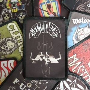 May include: A black and white embroidered patch with the text "Electric Wizard" in a curved font. The patch features a stylised image of a person lying on a platform with a sword piercing them. The patch has a black border.