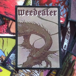 Op de afbeelding: Een zwart-wit geweven patch met de bandnaam "Weedeater" in gotische letter. De patch heeft een gedetailleerde illustratie van een draak op een textuur achtergrond.