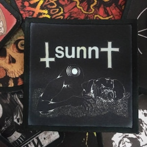 May include: Black and white embroidered patch with a cross and the word "sunn" in white text. A stylised image of a person lying down is in the bottom half of the patch.