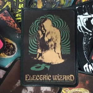 May include: A black rectangular patch with a beige seated figure and a green and teal swirl design. The gold text "ELECTRIC WIZARD" is below the figure. Other patches are visible in the background.