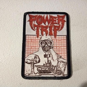 Könnte beinhalten: Ein gestickter schwarzer und weißer Aufnäher mit den Worten "POWER TRIP" in roten Buchstaben. Der Aufnäher zeigt eine Person mit Gasmaske und Schutzanzug, die vor einem roten und weißen Gittermuster steht.