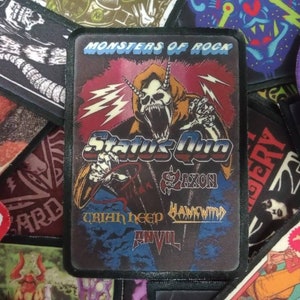 May include: A black and white patch with a red, blue and yellow design. The patch features a monster with horns and a lightning bolt. The text "Monsters of Rock" is at the top of the patch. The text "Status Quo" is in the centre of the patch. The text "Saxon" is below the monster. The text "Uriah Heep Hawkwind Anvil" is at the bottom of the patch.
