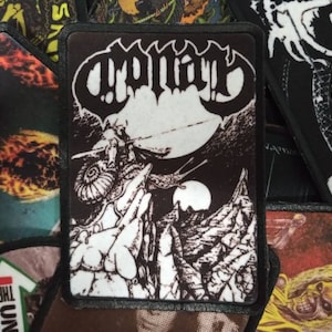 May include: Black and white embroidered patch with the band name "CRYPTIC" in a gothic font. The patch features a black and white illustration of a creature with a large eye and a long, thin body.