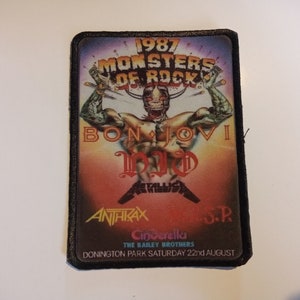 May include: A black and white patch with a colorful image of a muscular figure with a skull face. The text "1987 Monsters of Rock" is at the top of the patch. The text "Bon Jovi" is in the middle of the patch. The text "Anthrax" is at the bottom of the patch. The text "Donington Park Saturday 22nd August" is at the bottom of the patch.