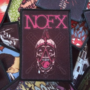 Könnte beinhalten: Ein schwarzer und pinkfarbener gestickter Patch mit dem Bandnamen "NOFX" in pinken Buchstaben. Der Patch zeigt einen Schädel mit einem pinkfarbenen Maulkorb und einem pinkfarbenen Lederhalsband. Zwei gekreuzte Paddel befinden sich hinter dem Schädel.