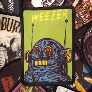 May include: An embroidered patch in green and black with the word "Weezer" in white lettering. The patch features a cartoon robot with blue and yellow details.