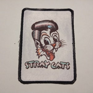 Può includere: Una toppa bianca con cuciture nere attorno ai bordi. La toppa presenta un gatto dei cartoni animati con un'acconciatura pompadour blu e bianca. Il gatto ha una lingua rosa che sporge. Il testo "Stray Cats" è stampato in un font arcobaleno sotto il gatto.