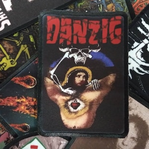 Könnte beinhalten: Schwarzer Aufnäher mit dem Wort "DANZIG" in Rot, einem Totenkopf und einer religiösen Figur mit einem leuchtenden Herzen. Weitere Aufnäher mit einem Skelett und einem Kometen sind sichtbar. Für Kleidung oder Accessoires.