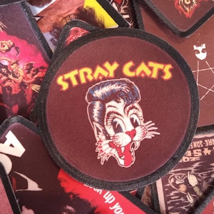 May include: A maroon fabric patch with a black border and white stitching. The patch features a cartoon cat with a red tongue sticking out and the text "Stray Cats" in yellow letters.
