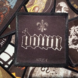 May include: A black and white embroidered patch with the word "DOWN" in a gothic font. The patch has a fleur-de-lis design above the word and the text "NOLA" below it.