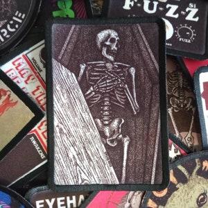 May include: A black and white embroidered patch featuring a skeleton in a coffin. The patch has a brown background and black stitching around the edges.