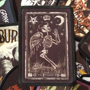 May include: A black and white tarot card patch featuring a skeleton angel with wings. The card is labelled "The Sinner" and has a crescent moon and a pentagram in the top right corner.