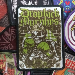 Könnte beinhalten: Ein gestickter Patch in Grün, Schwarz und Weiß mit dem Text "Dropkick Murphys" und einem Skelett, das Boxhandschuhe trägt.
