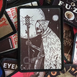May include: A black and white illustration of a skeletal figure in a hooded robe holding a lantern. The figure is standing in front of a large, full moon with stars in the background. The illustration is printed on a brown fabric patch.