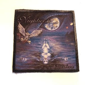 Peut inclure: Un patch brodé noir et blanc sur fond bleu foncé. Le patch représente une femme aux longs cheveux noirs flottant dans l'eau, un oiseau blanc avec un ruban rouge volant au-dessus d'elle, et un grand œil bleu et blanc dans le ciel. Le texte "Nightwish" et "Oceanborn" est visible sur le patch.