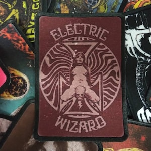 May include: A red and black embroidered patch with the text "Electric Wizard" in white lettering. The patch features a stylized image of a woman with a skull in her lap.