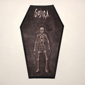 Puede incluir: Un parche bordado en blanco y negro con forma de ataúd con la palabra "GOJIRA" en la parte superior y un esqueleto en su interior.