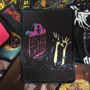 Peut inclure: Patch brodé noir avec une pierre tombale rose et bleue sur laquelle est inscrit "Electric Wizard". Une main squelettique sort du sol.