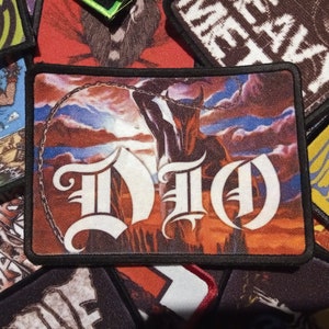 May include: A black and white embroidered patch with the word "DIO" in a gothic font. The patch features a red, orange, and blue background with a silhouette of a figure in the background.