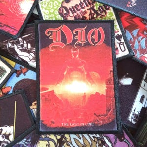 May include: A black and white embroidered patch featuring the band name "DIO" in a gothic font. The patch has a red background with a silhouette of a figure wearing a horned helmet. The text "The Last in Line" is printed below the figure.