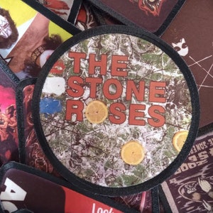 Puede incluir: Un parche redondo con borde negro y fondo de camuflaje. El texto "THE STONE ROSES" está impreso en letras rojas y blancas.