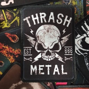 May include: Black and white embroidered patch with a skull and crossbones design. The text "THRASH METAL" is written in a bold font with the year "666" and "EST" on either side of the skull.