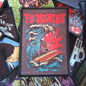 Peut inclure: Un écusson brodé noir et marron avec le texte "PRESENTING FU MANCHU" en blanc. L'écusson représente un squelette sur une planche de surf rouge dans une vague bleue avec un soleil rouge en arrière-plan.