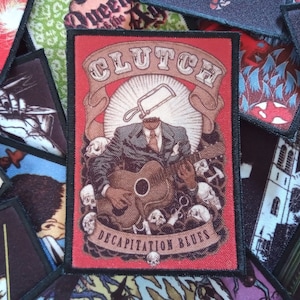 May include: A red, white and black woven patch with the band name "CLUTCH" in white lettering and the words "DECAPITATION BLUES" in white lettering on a banner. The patch features a man in a suit with a saw for a head playing a guitar.