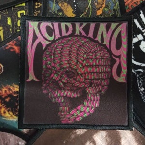 May include: A black and white patch with the text "Acid King" in pink and green letters. The patch features a skull with a pink and green pattern.
