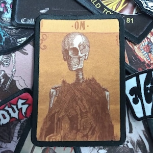 May include: A black and white embroidered patch featuring a skeleton wearing a brown coat. The patch has a yellow background and the text "OM" in the top right corner.
