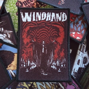 May include: A black and red embroidered patch with the text "WINDHAND" in white lettering. The patch features a figure in a black cloak standing in front of a red and black background.