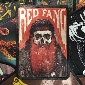 Peut inclure: Patch de groupe Red Fang avec un crâne et une barbe dans un schéma de couleurs rouge et noir. Le patch a une bordure noire et le nom du groupe est écrit en lettres blanches.