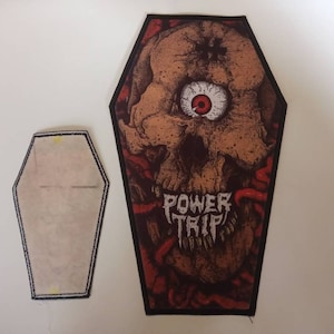 May include: A black and red coffin-shaped patch featuring a skull with one eye and the text "POWER TRIP" in white.