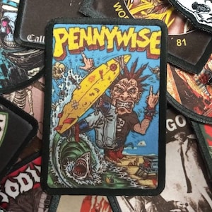 May include: A black and yellow embroidered patch with the band name "PENNYWISE" in white letters. The patch features a cartoon image of a punk rocker riding a surfboard with a shark in the water below.
