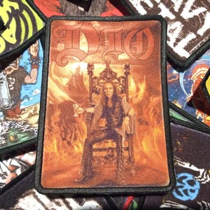 May include: A black and white embroidered patch with the word "DIO" in a stylised font. The patch features a depiction of a man sitting on a throne in front of a fiery background.