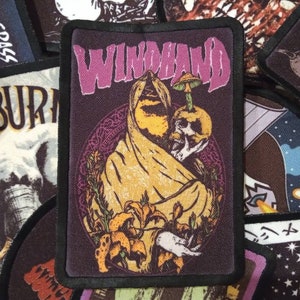 May include: A black and purple embroidered patch with the word "WINDHAND" in purple lettering. The patch features a hooded figure holding a mushroom and a skull with flowers in the background.
